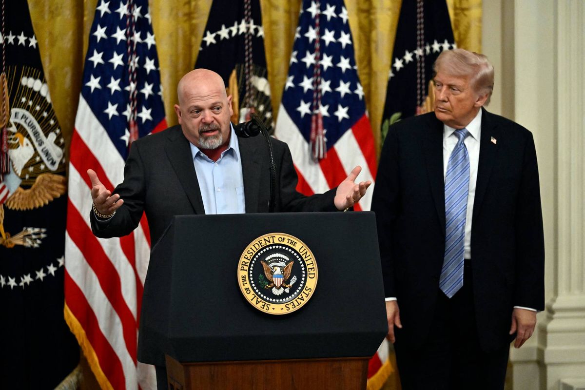 Two men in formal attire are standing at a podium, one speaking and the other listening. Behind them are several American flags and a backdrop that suggests a formal setting, likely a press conference or official announcement.
