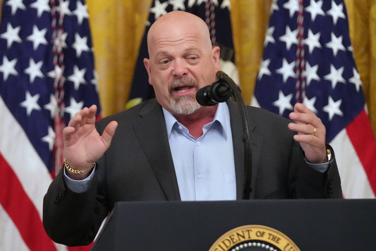 A man in formal attire, standing before a microphone, appears to be delivering a speech. In the background, American flags are prominently displayed, suggesting a formal or political setting.