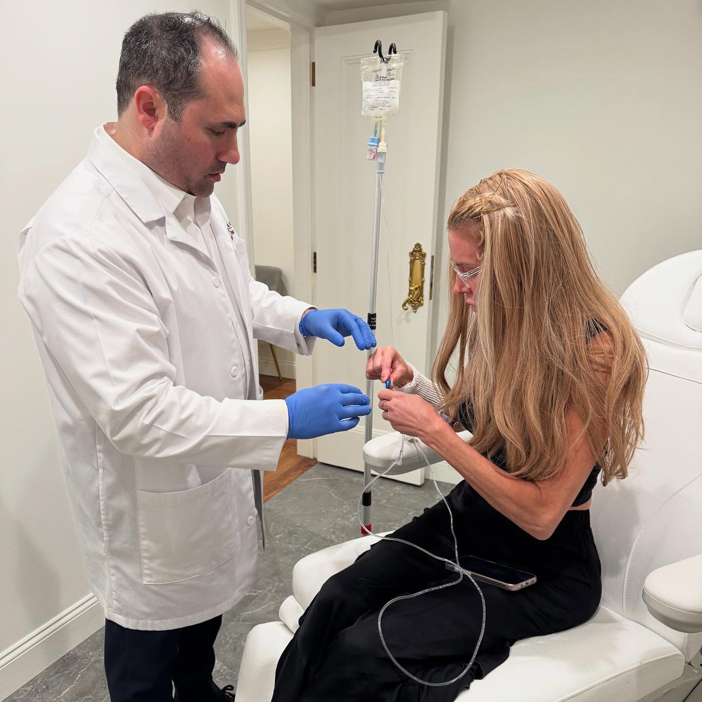 A doctor in a white coat and blue gloves prepares an IV drip for a woman with long blonde hair, who is seated and holding part of the IV line.