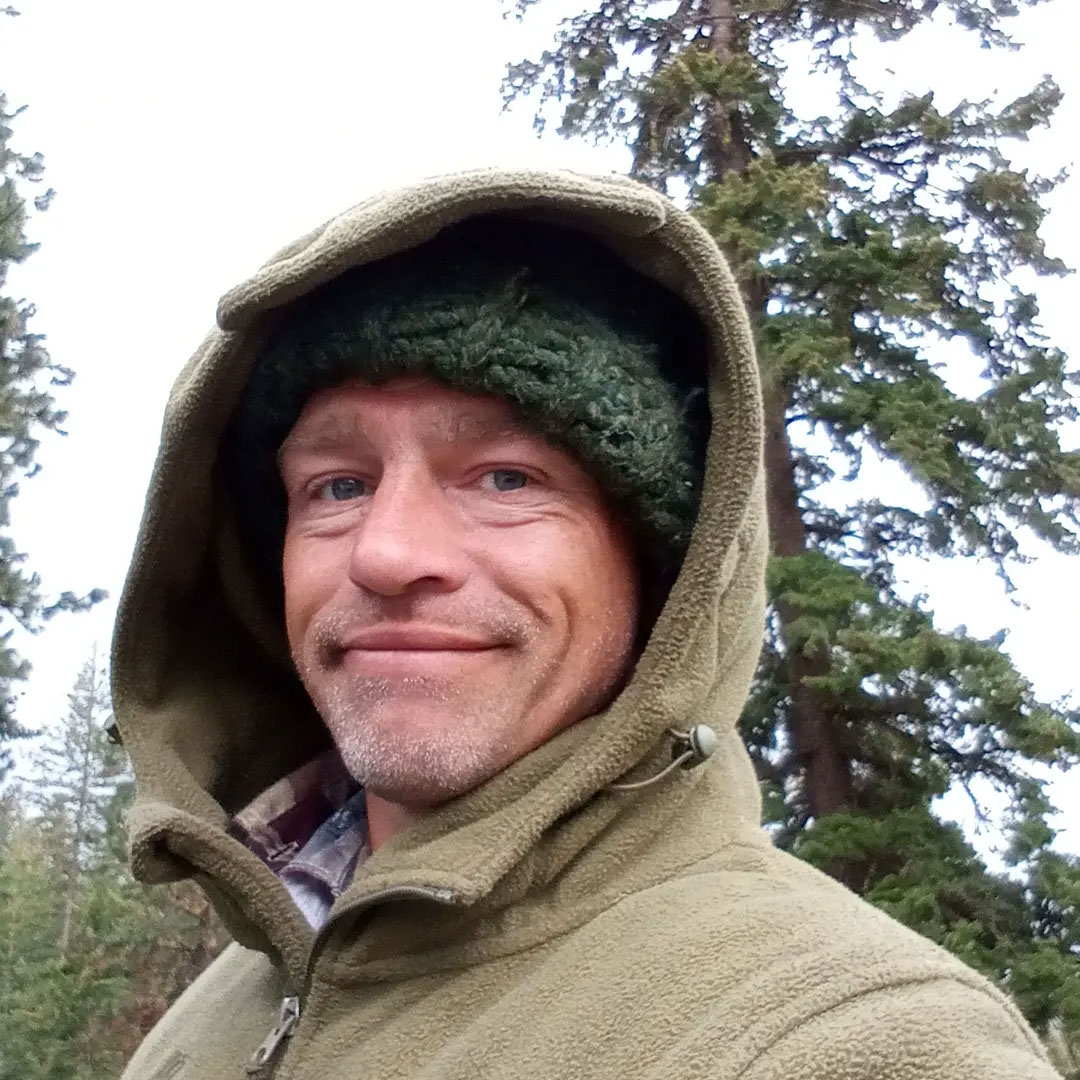 Matt Brown, wearing a hooded jacket and knit cap, smiling outdoors with trees in the background.