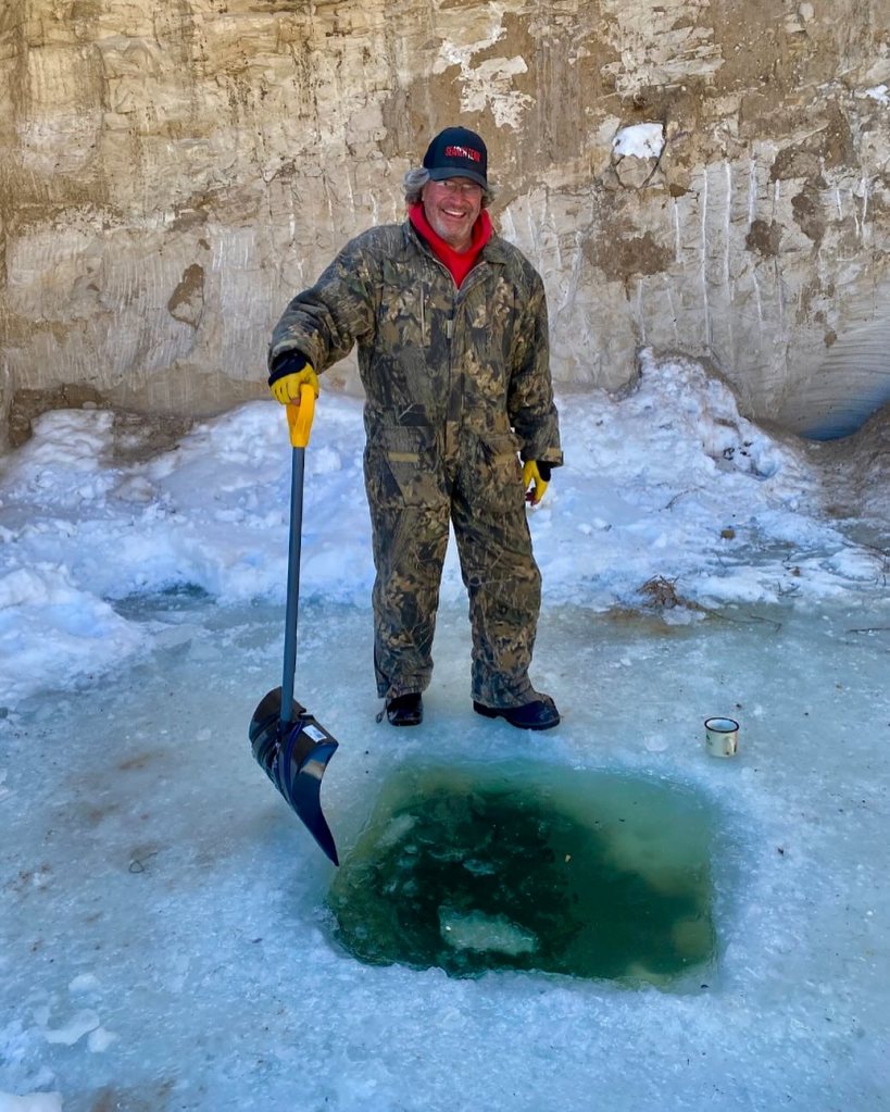 Duane Ollinger standing on ice with a shovel next to a hole in the ice.