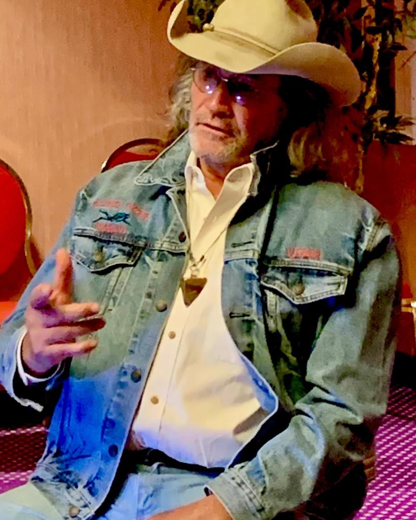 Duane Ollinger wearing a cowboy hat, glasses, and a denim jacket, sitting and gesturing with his right hand.