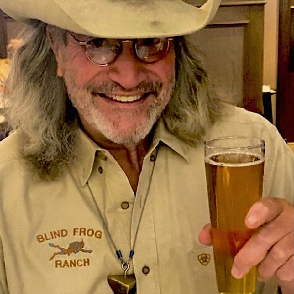 Duane Ollinger wearing a cowboy hat and a "Blind Frog Ranch" shirt, holding a glass of beer.