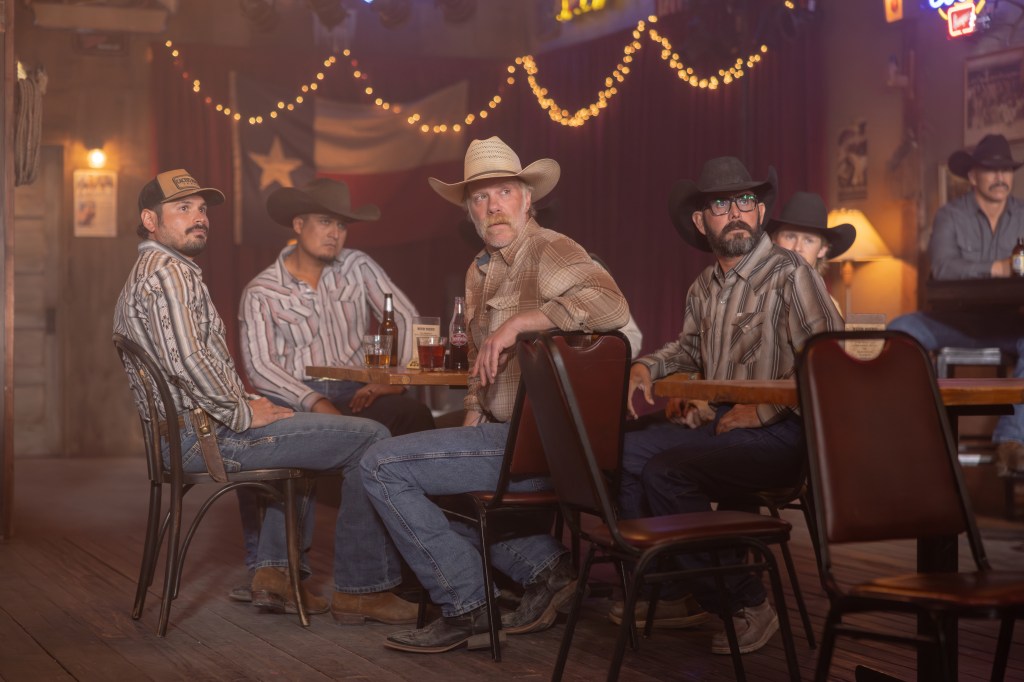 Azul and Zachariah sit at a table in a bar with other cowboys.