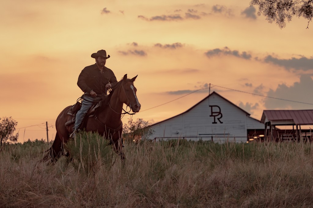 Cole Hauser as Rip Wheeler on horseback in front of a barn with "DR" on it.