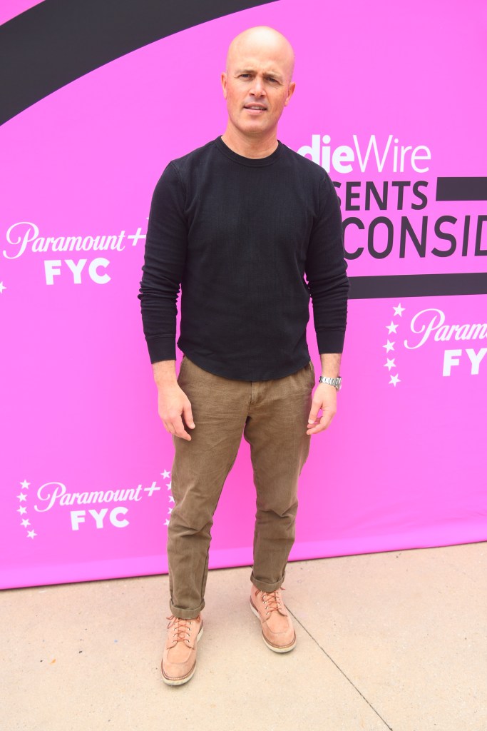 Chad Feehan in a black long-sleeved shirt and brown pants posing in front of a pink backdrop.